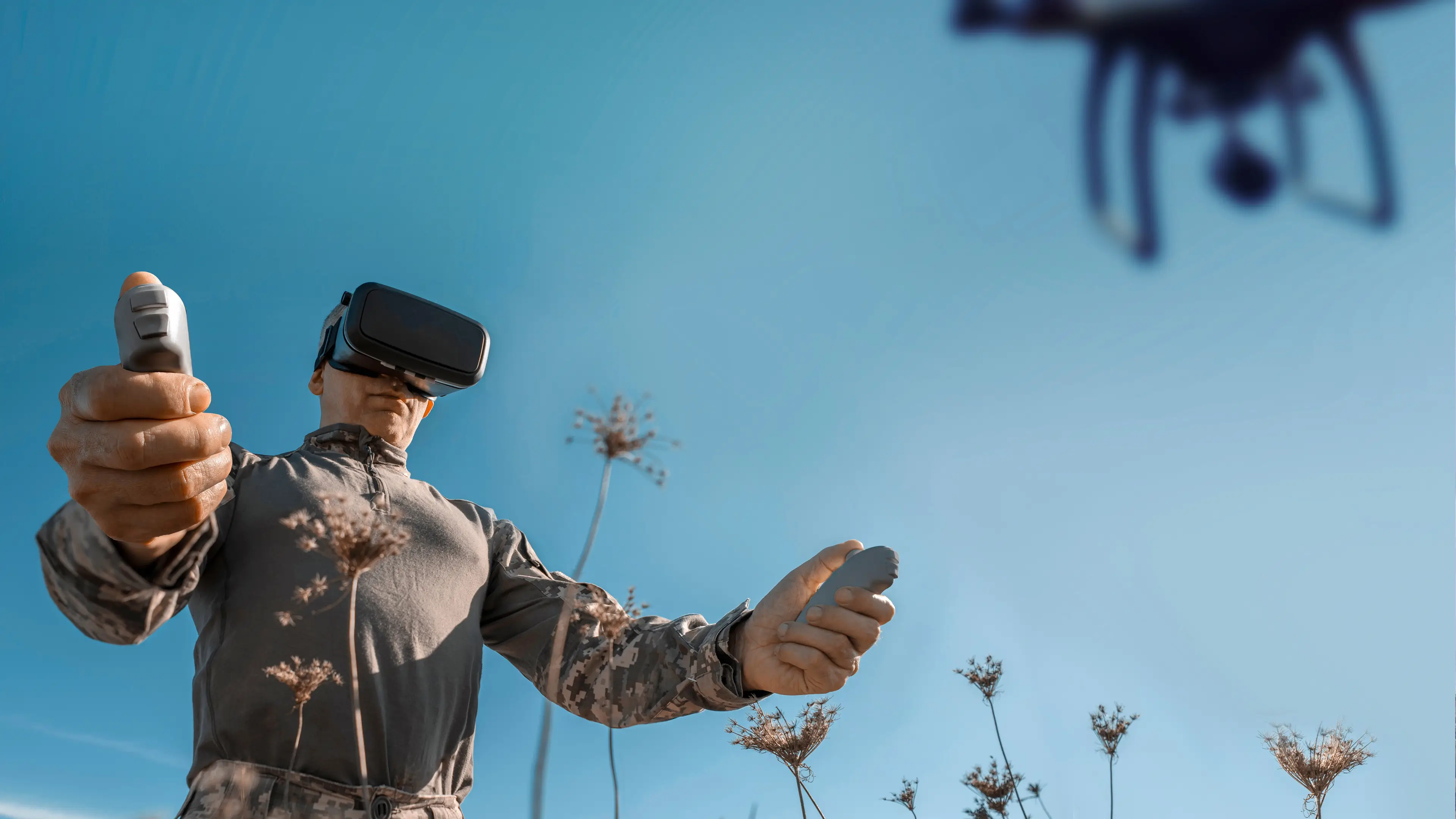 A person wearing a virtual reality headset and camouflage clothing is outdoors, holding two handheld controllers. Tall dried plants are visible in the foreground, and a drone is flying in the sky above against a clear blue background.   Image depicting the Defence market