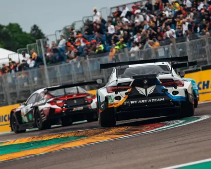 Two GT race cars speeding on a track with colorful curbs, viewed from behind, as a crowd watches from grandstands lined with Michelin banners.