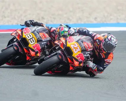 Two MotoGP riders leaning sharply into a corner on a racetrack, riding orange and black motorcycles with visible numbers 33 and 23.