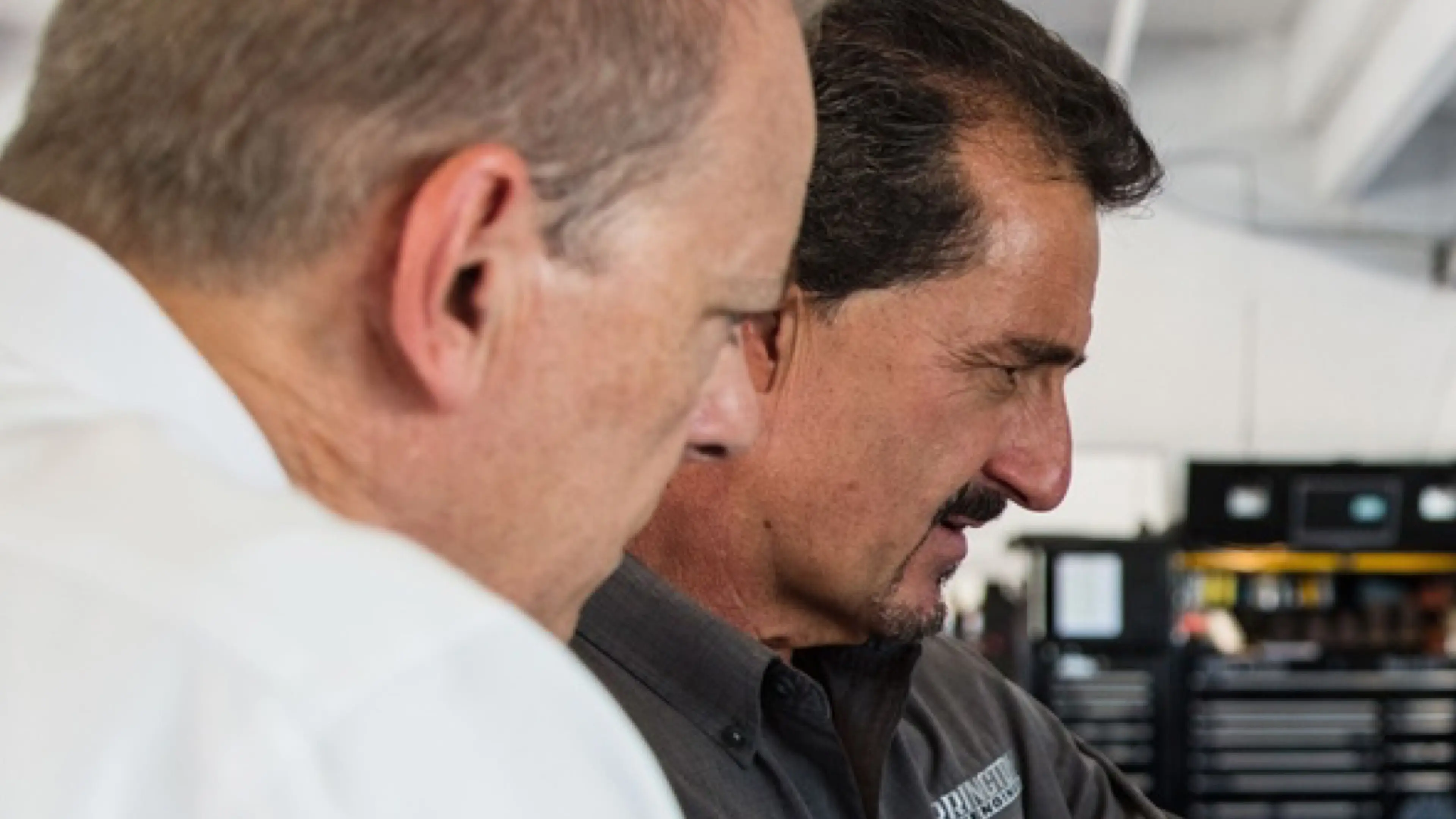 Two people wearing white “Motion Applied” shirts stand on a pit lane at a racetrack, with race cars, crew members, and a large colorful number sign overhead. In the background, uniformed individuals line up near the track and grandstands filled with spectators.