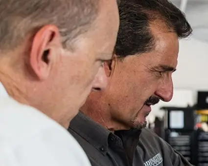 Two people wearing white “Motion Applied” shirts stand on a pit lane at a racetrack, with race cars, crew members, and a large colorful number sign overhead. In the background, uniformed individuals line up near the track and grandstands filled with spectators.