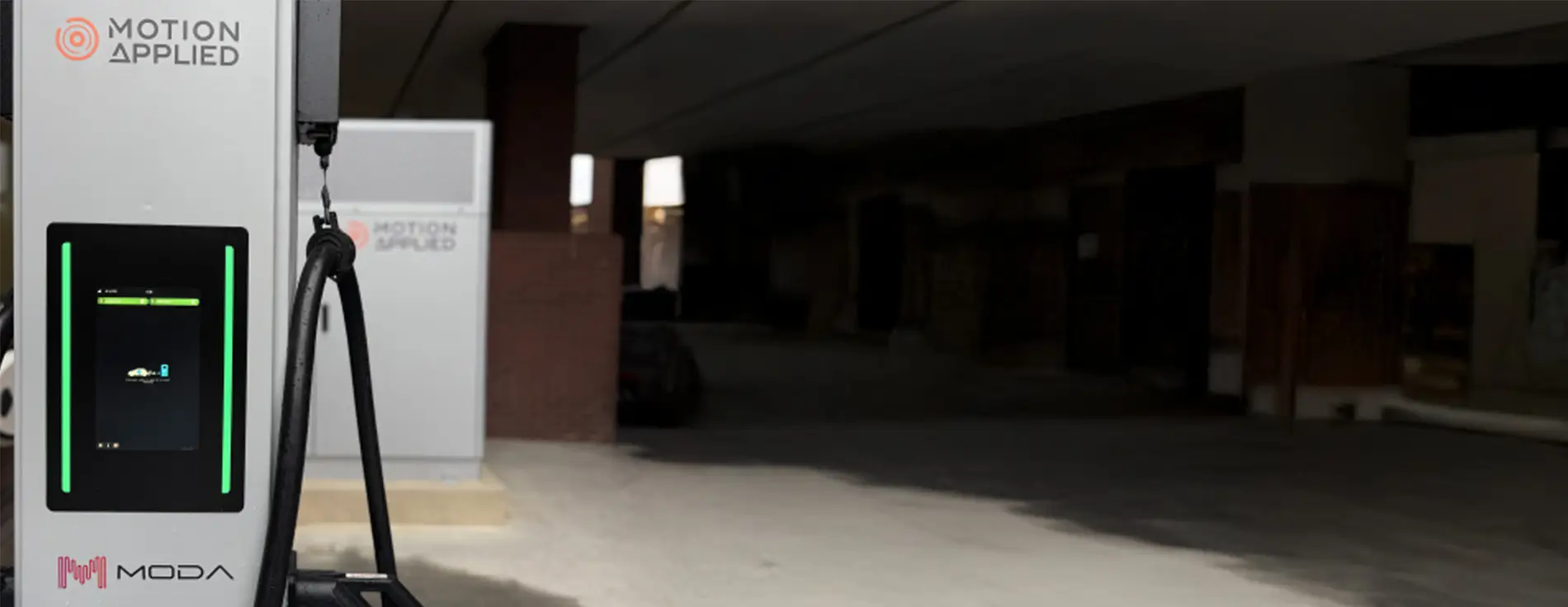 A sleek black electric car parked next to a white ultra-rapid EV charging station branded with “MODA” and “Motion Applied.” The charger has a digital display and pink cable graphics at the bottom. The setting is an urban area with a brick wall and modern office building in the background.