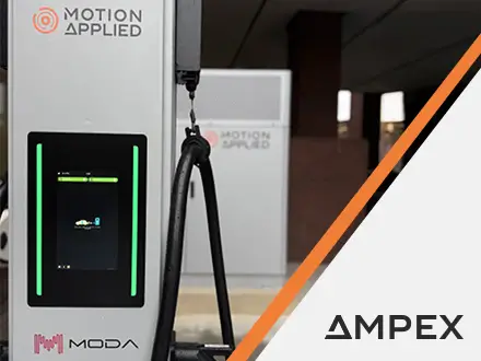 A sleek black electric car parked next to a white ultra-rapid EV charging station branded with “MODA” and “Motion Applied.” The charger has a digital display and pink cable graphics at the bottom. The setting is an urban area with a brick wall and modern office building in the background.