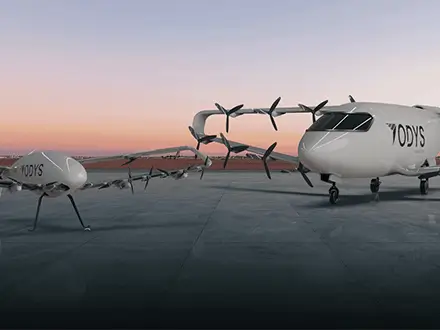 Two white electric aircraft with multiple rotors, branded ‘ODYS,’ positioned inside a hangar at sunset, with one larger aircraft and a smaller companion vehicle shown side by side.