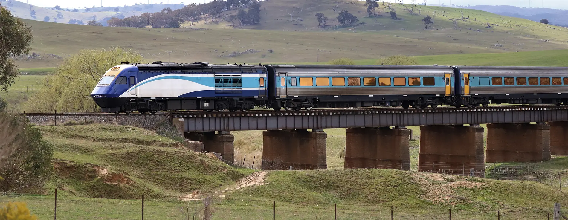 Blue and silver passenger train crossing a steel bridge over a rural valley, with rolling green hills, scattered trees, and farmland in the background.