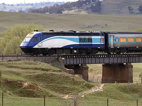 Blue and silver passenger train crossing a steel bridge over a rural valley, with rolling green hills, scattered trees, and farmland in the background.