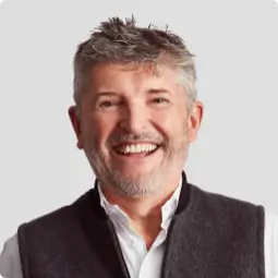 Nick Fry, Non-Executive Chairman, middle-aged with short styled light brown hair, smiling, wearing a white shirt undone at the top and a grey Gilet style jacket.