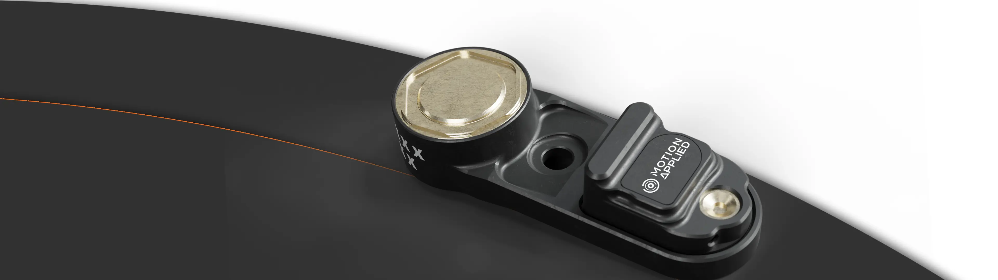 Compact Motion Applied electronic sensor module mounted on a black bracket, featuring a circular metal cap, a small rectangular housing with Motion Applied branding, and a mounting hole, shown against a dark background.