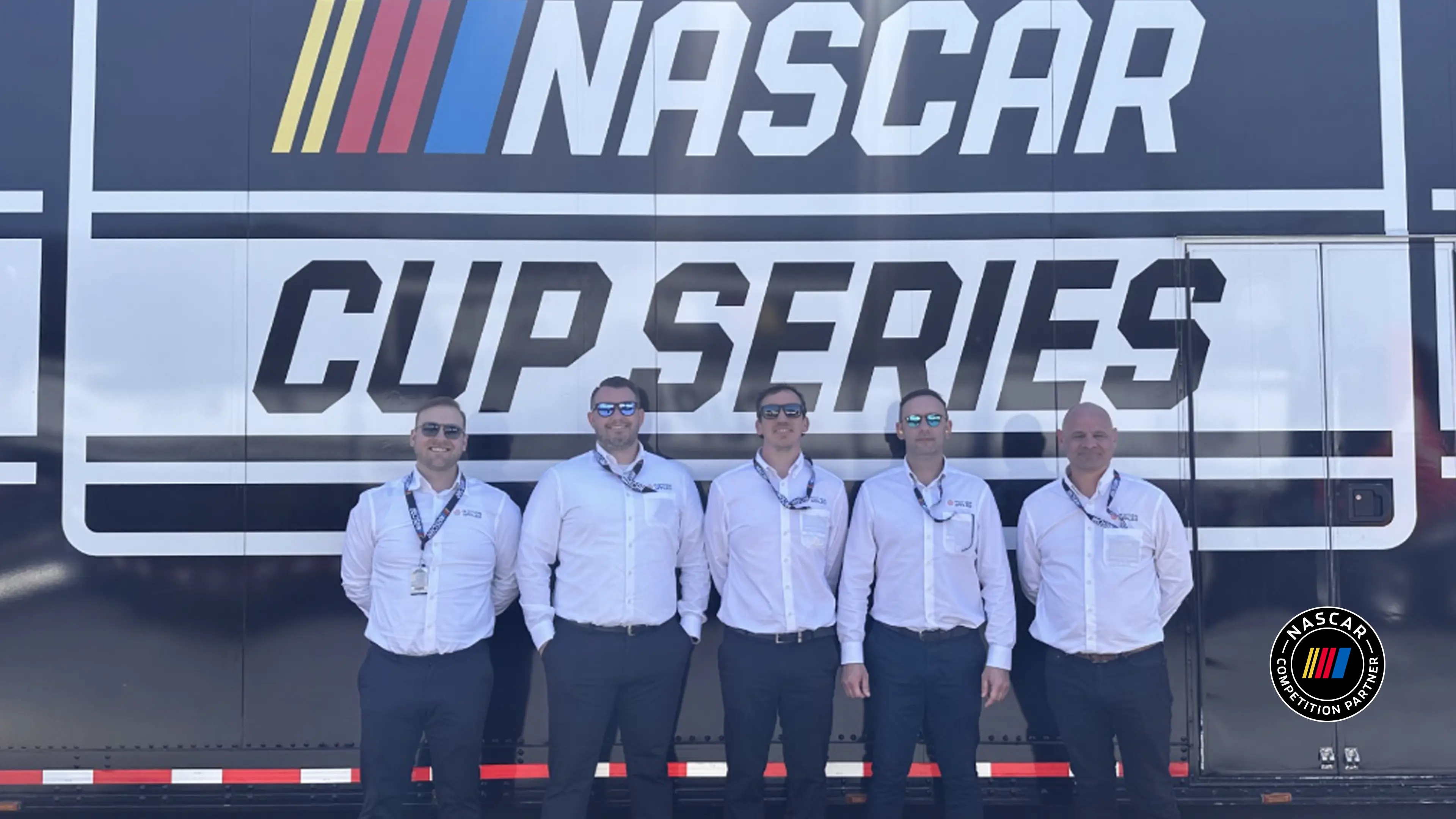 Five people wearing white shirts, stood in front of NASCAR signage